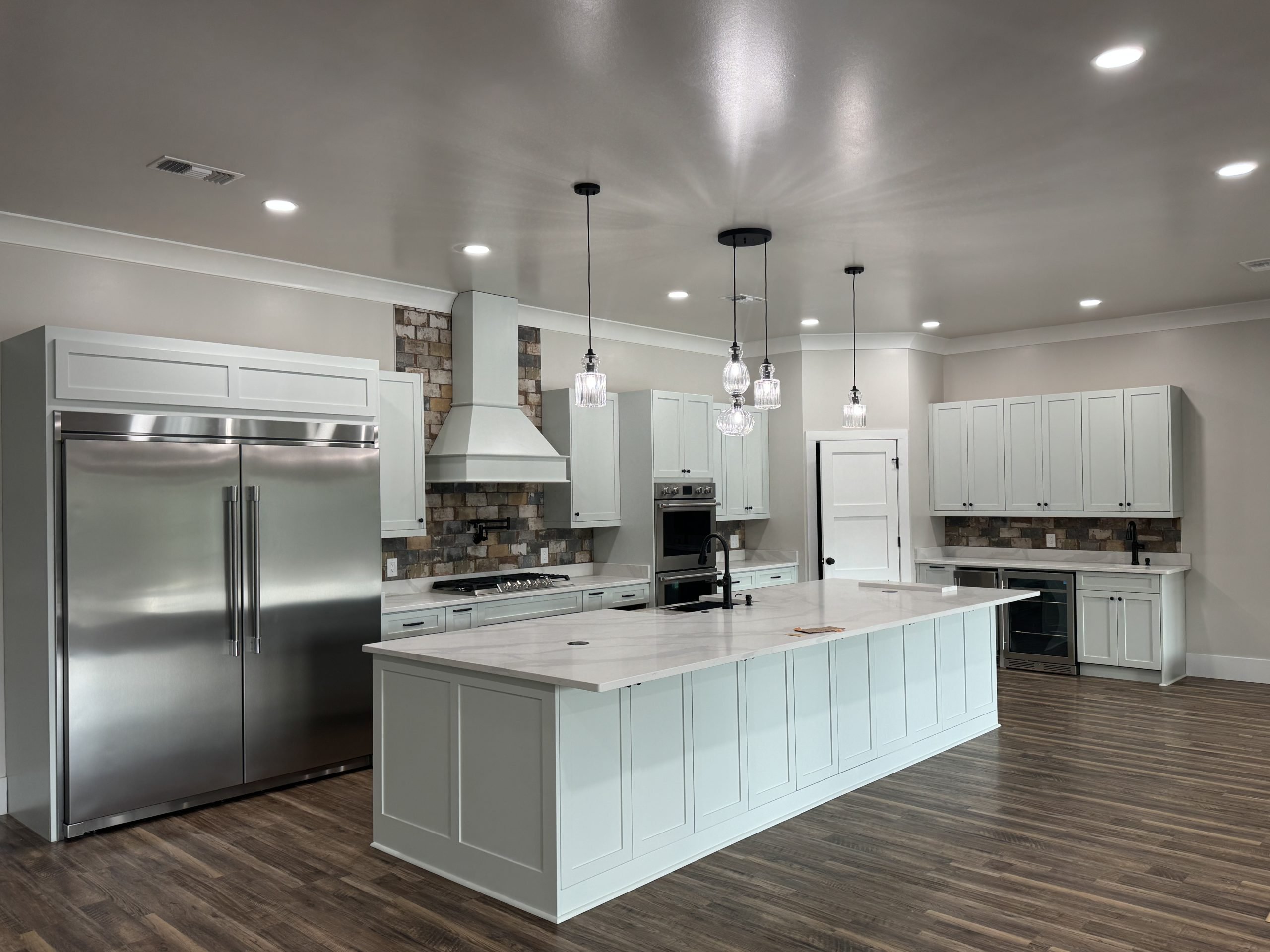 A spacious modern kitchen featuring white cabinetry, a large marble island with pendant lights above, stainless-steel double-door refrigerator, wall oven, and wood-style flooring.